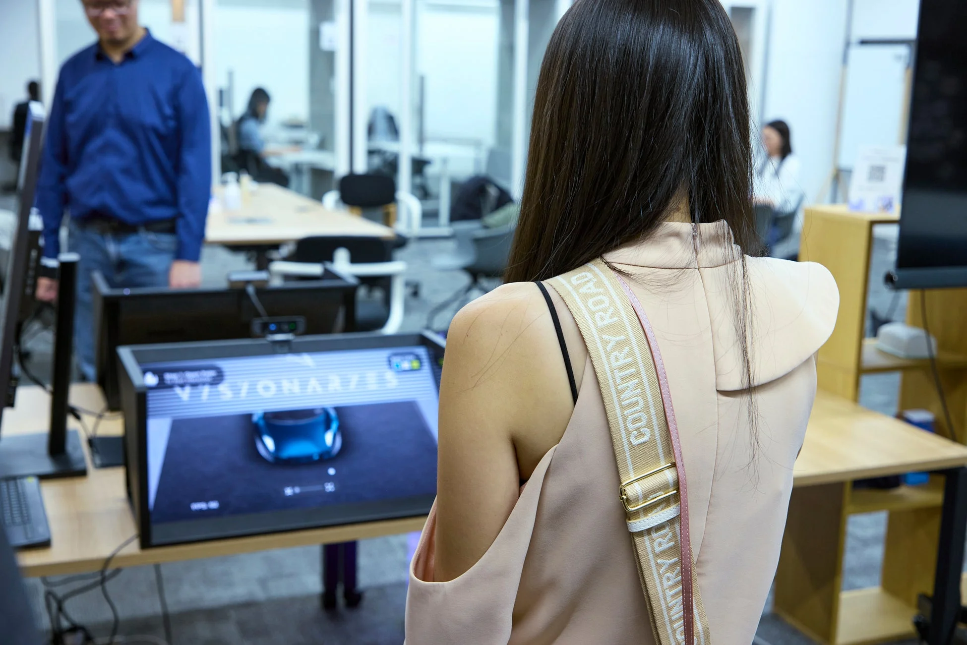 オフィスのような空間で、女性がディスプレイに映る青い車のデジタルモデルを見ている