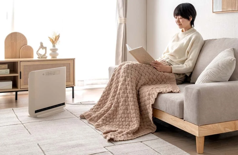 ソファでブランケットにくるまり読書を楽しむ女性と、足元に置かれたヒーターが写っています。暖かくリラックスしたリビングの様子を捉えた一枚です。