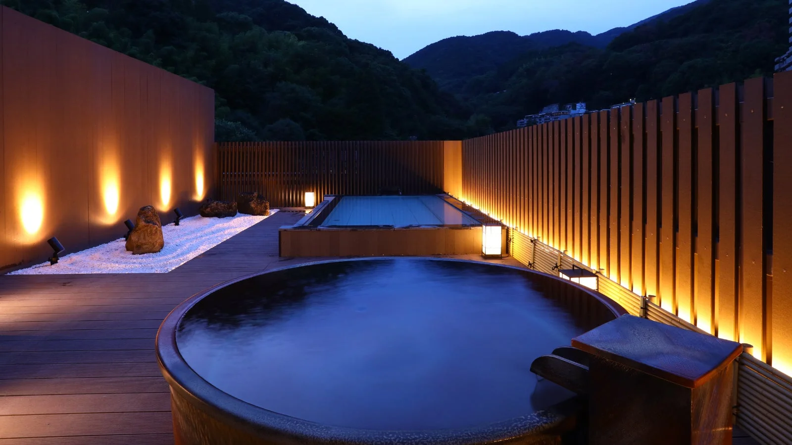 夕暮れ時、山々を背景にした露天風呂の風景。温かい光に照らされた円形と長方形の二つの湯船が、落ち着いた和の空間を演出し、リラックスできる雰囲気が漂っています。