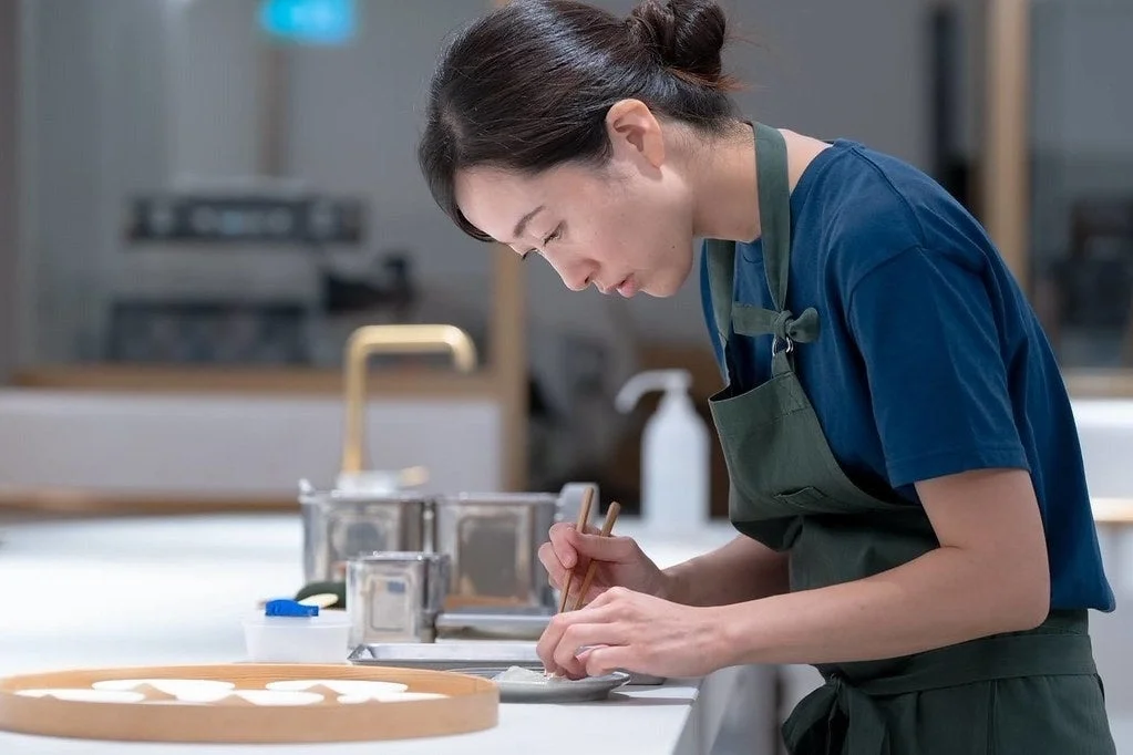 青いTシャツと緑のエプロンを着用した女性が、白いカウンターで箸を使い、小さな白い食材を丁寧に扱っています