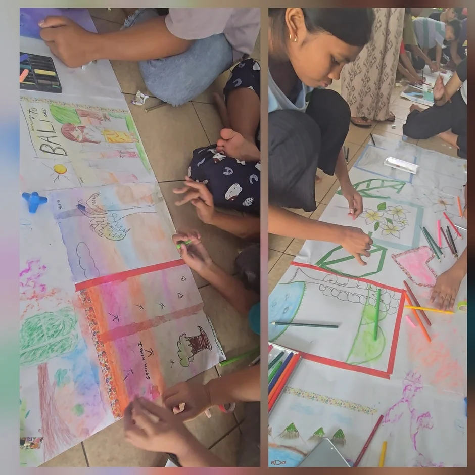 人々が床に座り、大きな紙に絵を描いている様子です。色鉛筆やクレヨンを使い、風景や花、文字など様々な絵が描かれており、共同でのアート活動が行われているようです。