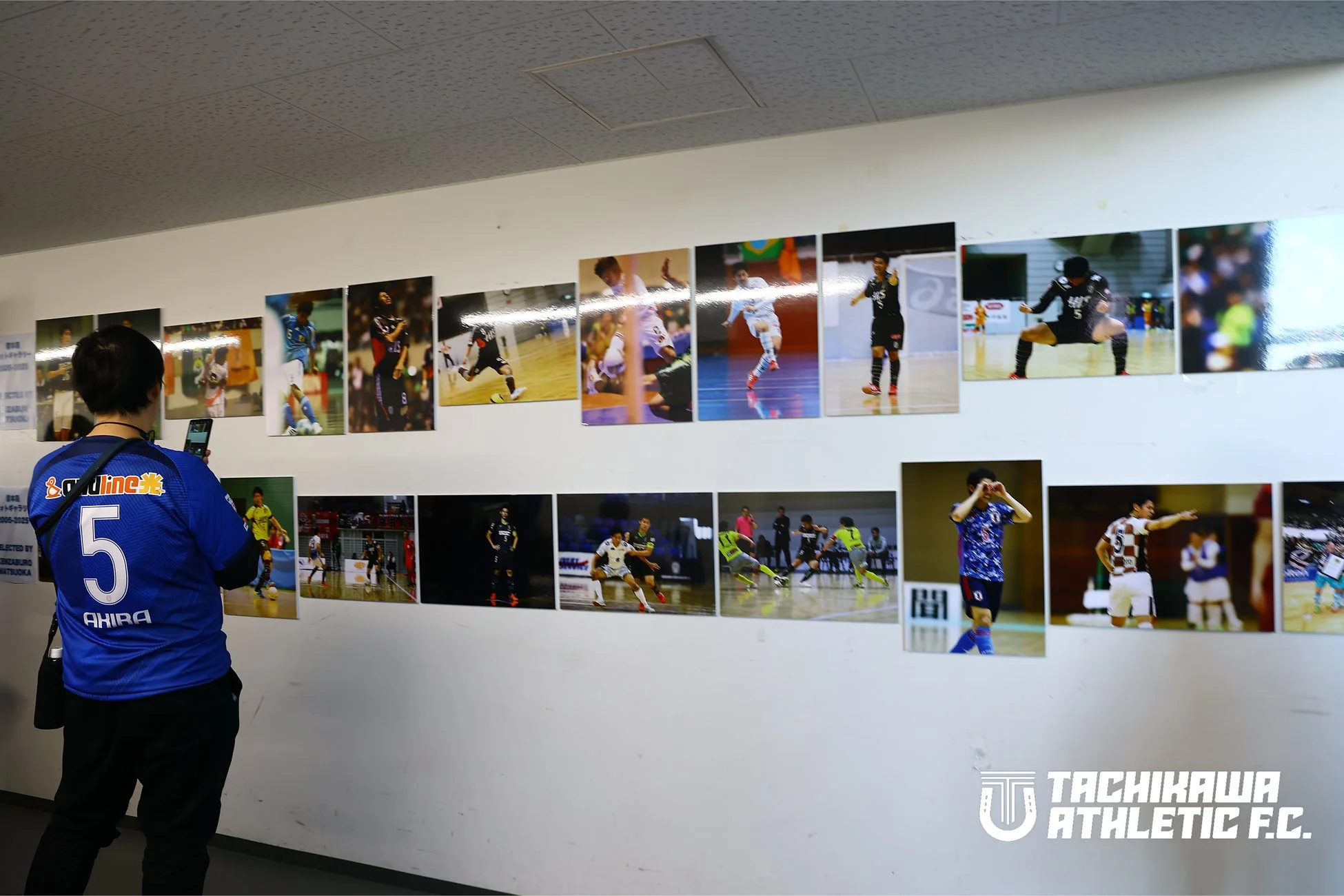 フットサル選手の躍動感あふれる写真が多数展示された壁を、背番号5の青いユニフォームを着た男性がスマートフォンで撮影している
