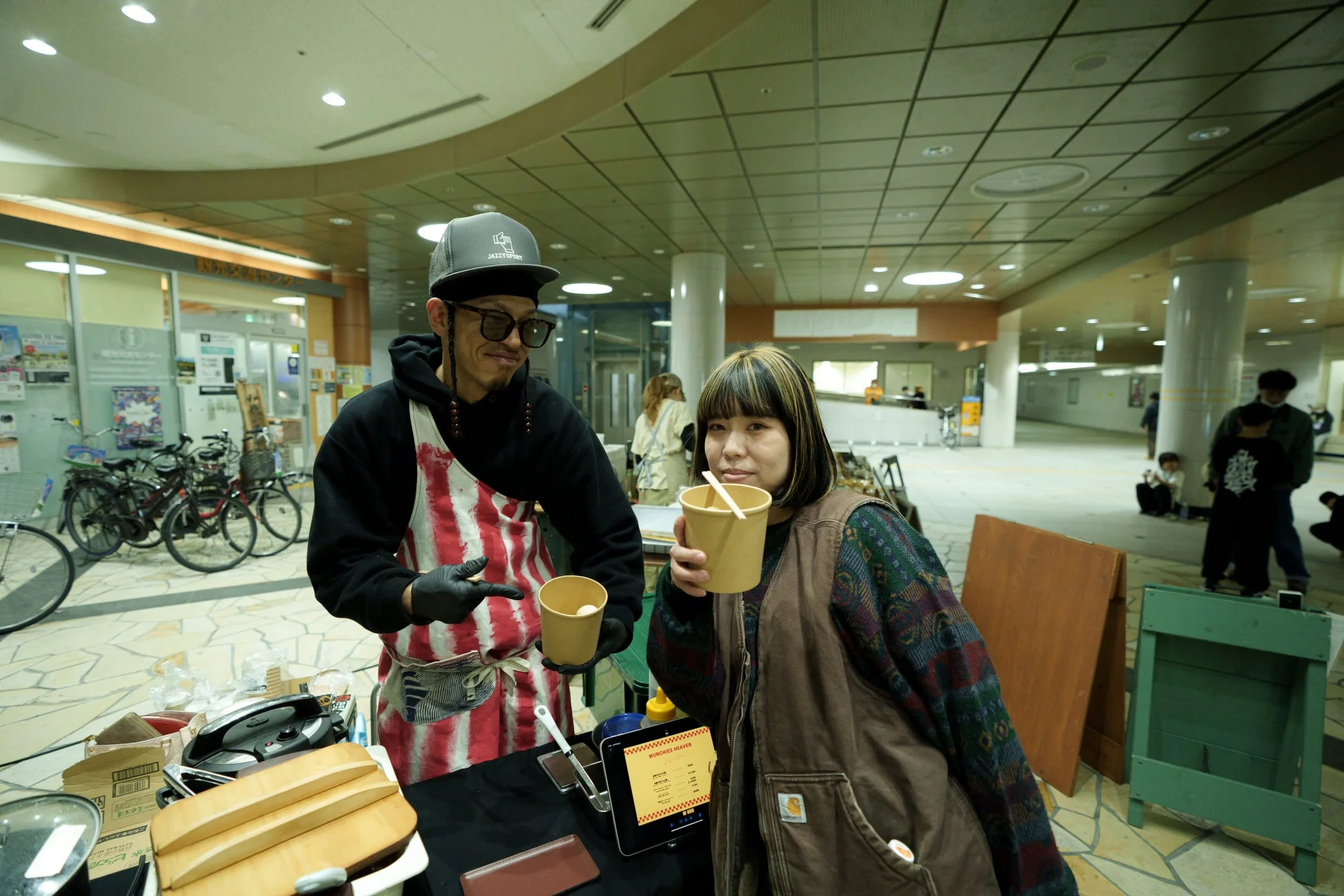 屋内イベントで、エプロンをつけた男性が飲み物を提供し、女性客がカップを手にしている様子