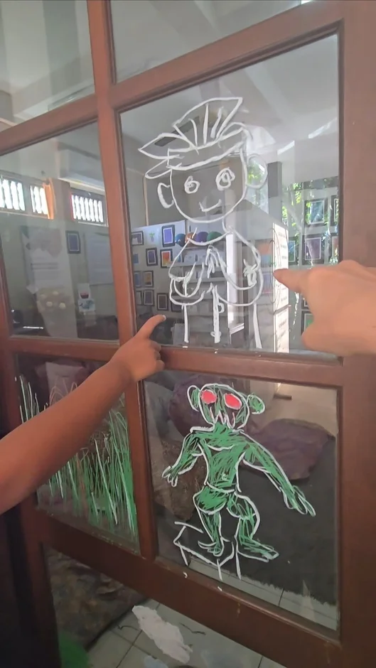 ガラス戸に描かれた白いキャラクターと緑のクリーチャーの絵を、二本の手が指差している。室内で展示されている手描きのイラストアートのようだ。