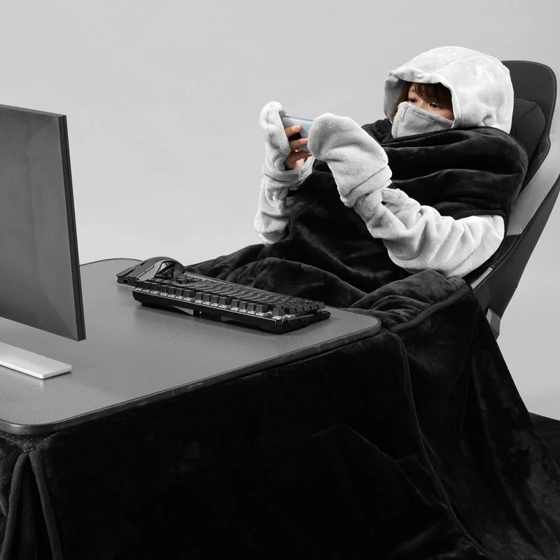 フード付きブランケットを着用した人物がゲーミングチェアに座り、スマートフォンを操作しています。デスクにはモニターやキーボードがあり、暖かくリラックスした雰囲気でゲームかSNSを楽しんでいるようです。