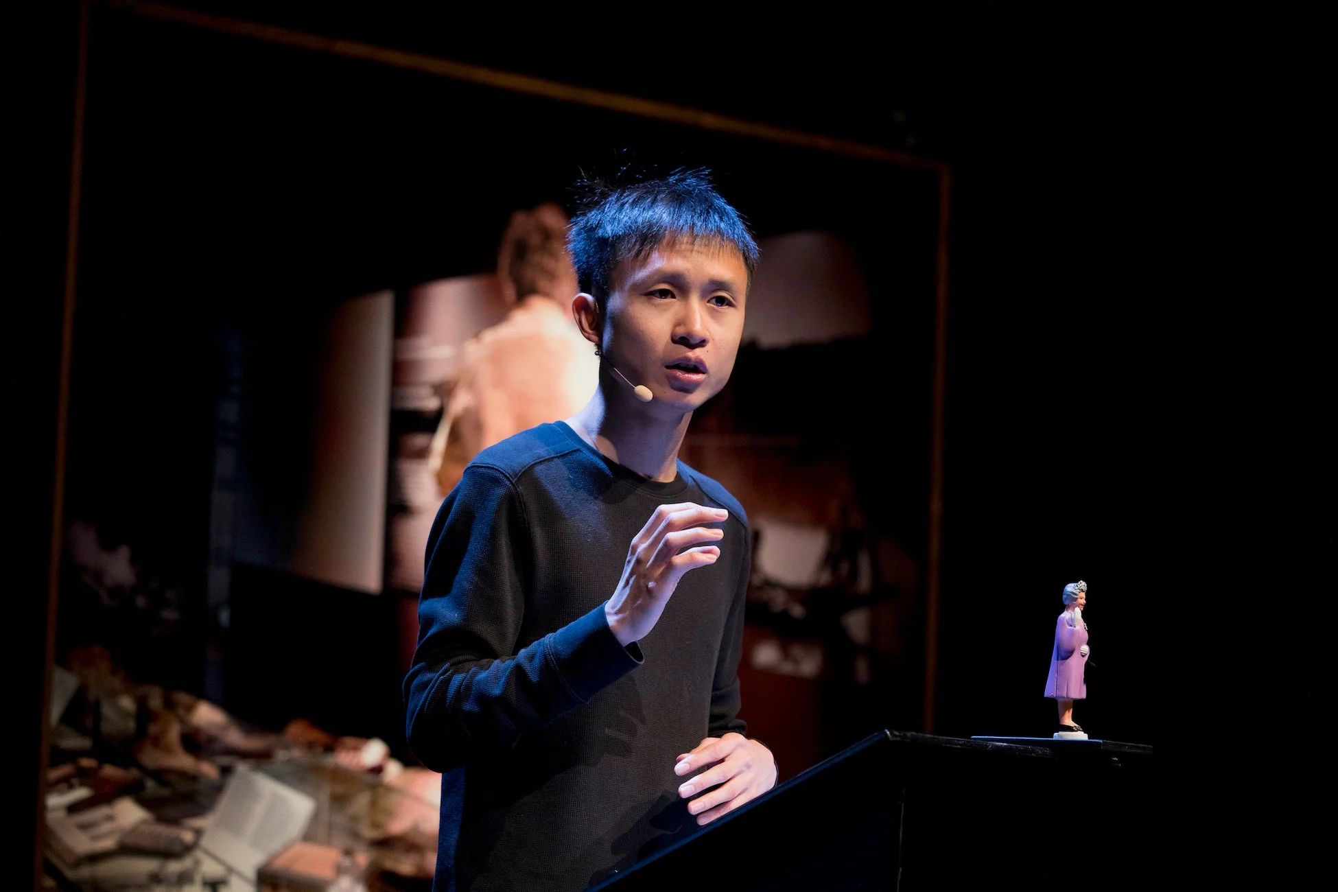 暗いステージ上で、ラベリアマイクを装着したアジア系の男性が熱心に話している