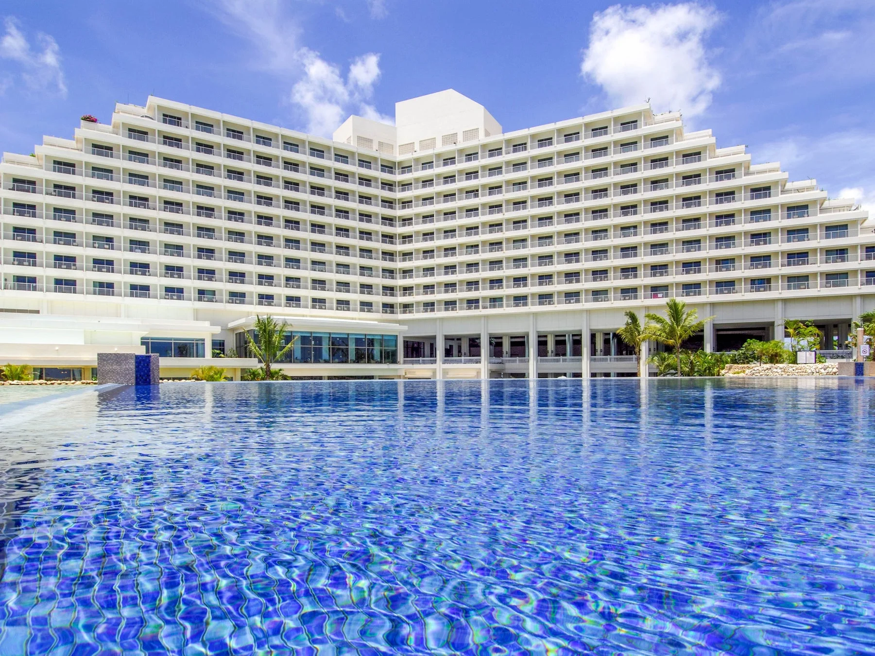 青い空と白い雲の下、広々とした屋外プールが手前に広がり、その奥にはテラスが連なる大規模な白いリゾートホテルが建っています。