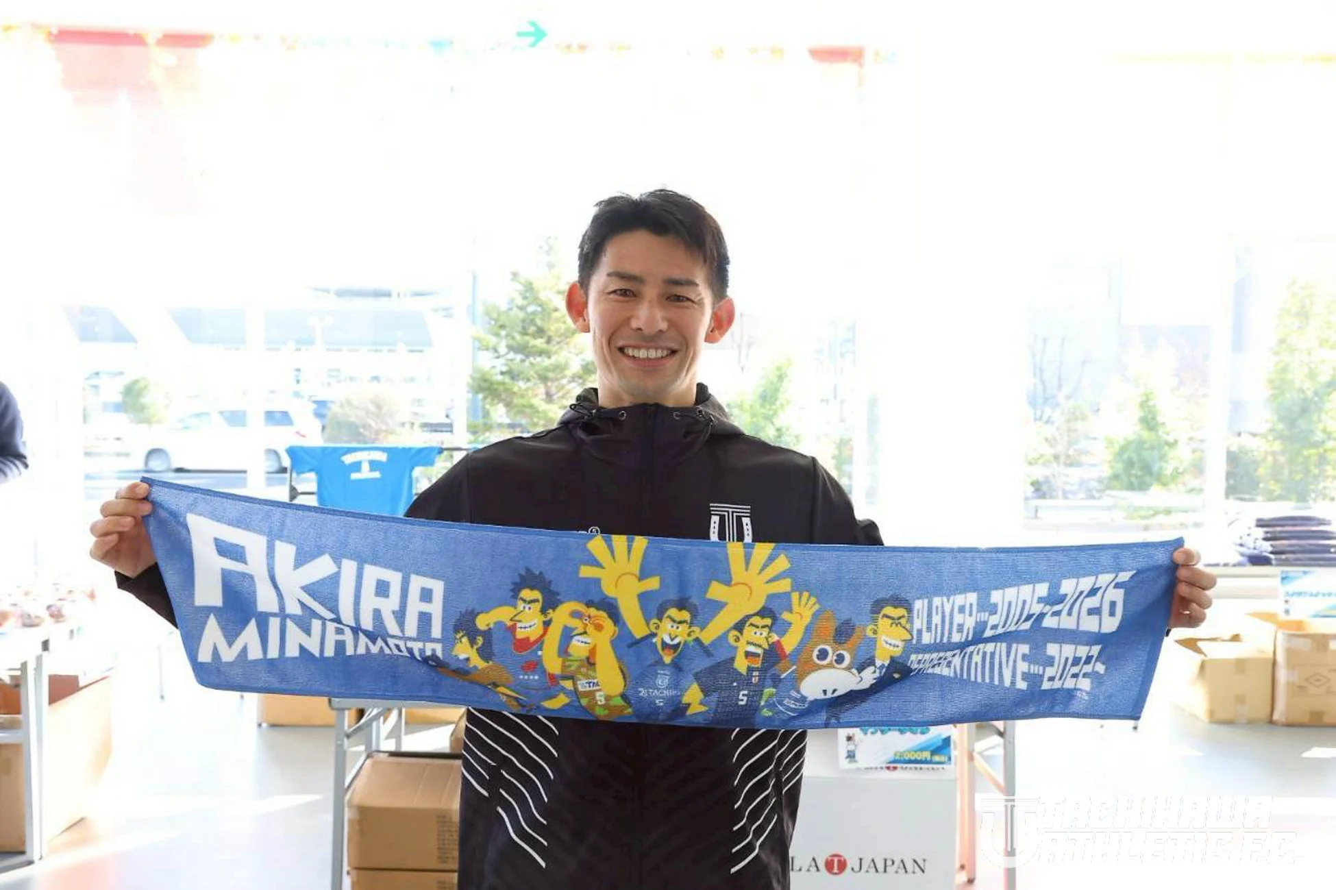 笑顔の男性が「AKIRA MINAMOTO」と書かれた青いタオルを両手で広げている