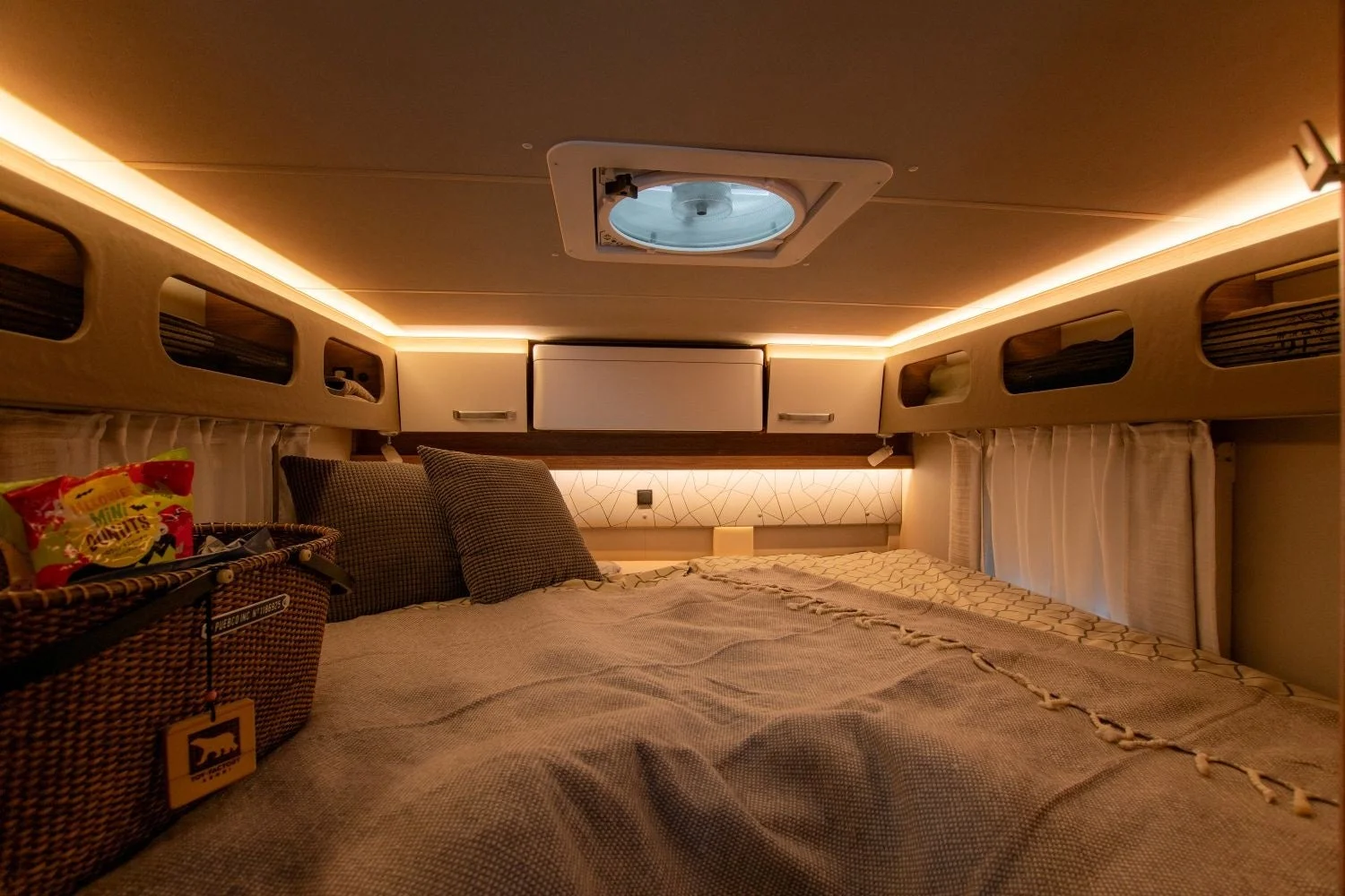 暖色系の間接照明が特徴的なキャンピングカーの寝室です。快適そうなベッドと枕があり、傍らにはスナックが入ったバスケットが置かれ、居心地の良い旅の雰囲気を醸し出しています。
