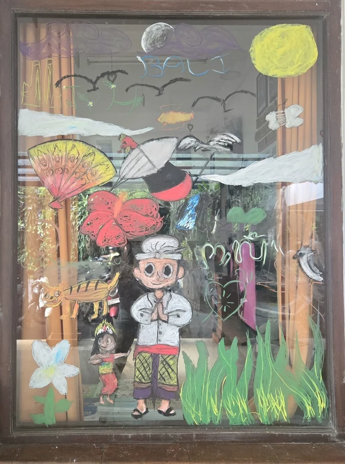 窓にチョークで描かれたバリ島の文化をテーマにしたカラフルなアート作品。伝統衣装を着た人物、動物、自然のモチーフが描かれ、「BALI」の文字も見られます。子供らしい素朴なタッチが特徴です。