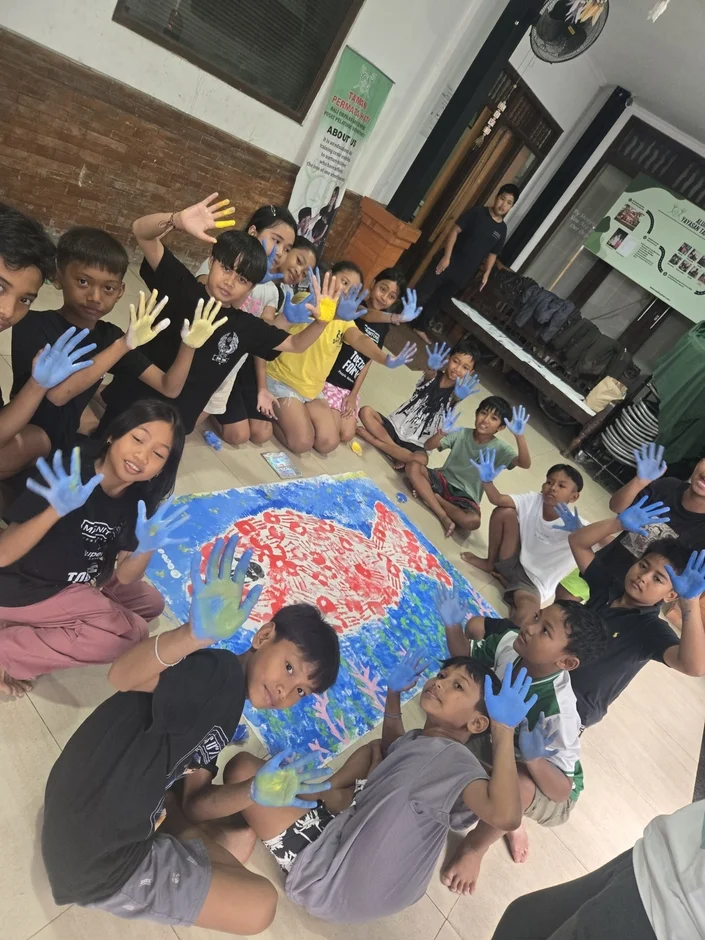 多くの子どもたちが床に座り、手形を使った大きな絵画を制作しています。彼らの手は青や黄色の絵の具で覆われ、笑顔で手を上げています。孤児院または訓練センターで行われているアート活動のようです。