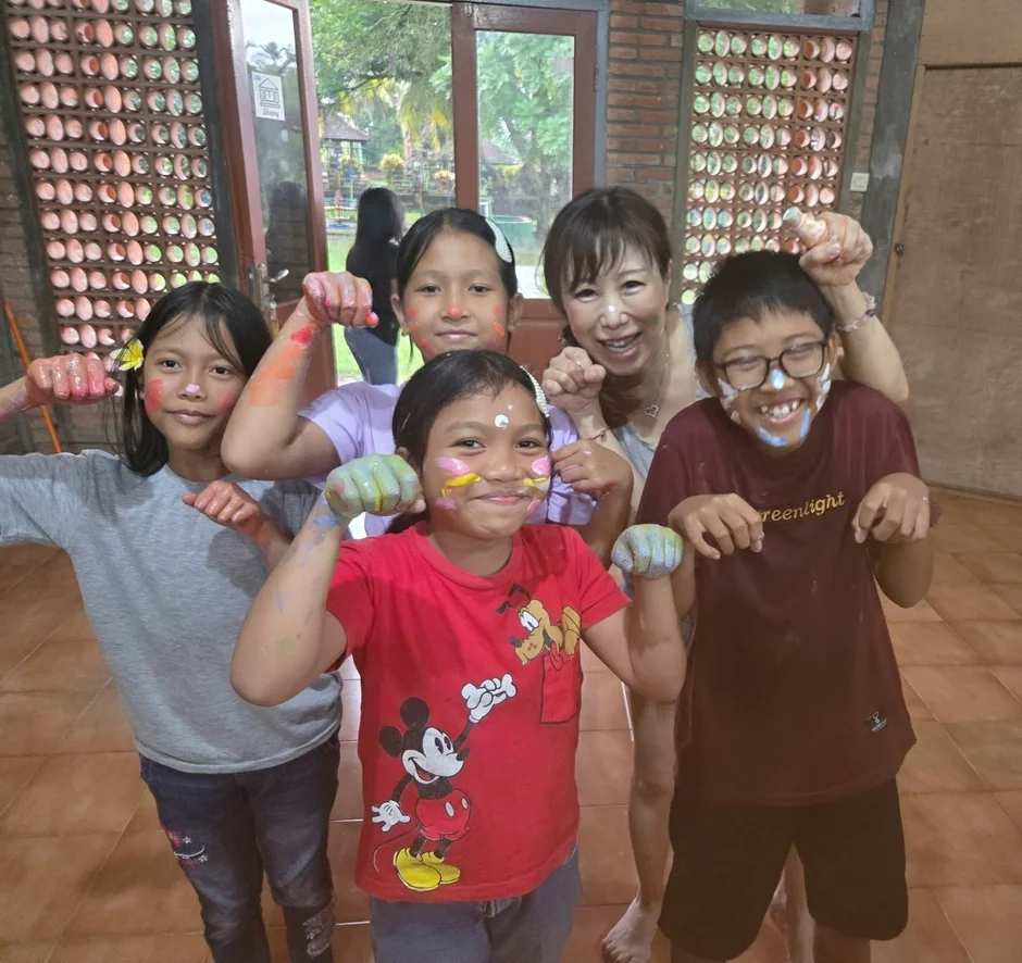 子供たちと大人の女性が、顔や手にカラフルな絵の具を塗って楽しんでいる様子を捉えた写真です。全員が笑顔でポーズをとっており、楽しい時間を過ごしていることが伝わります。