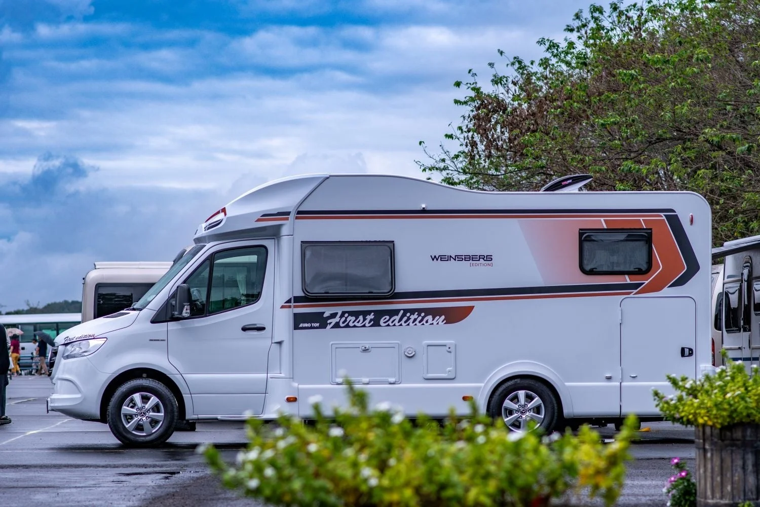 白い「WEINSBERG First edition」キャンピングカーが屋外に駐車されています。車体側面にはオレンジと黒のストライプがあり、背景には曇り空と木々が見えます。