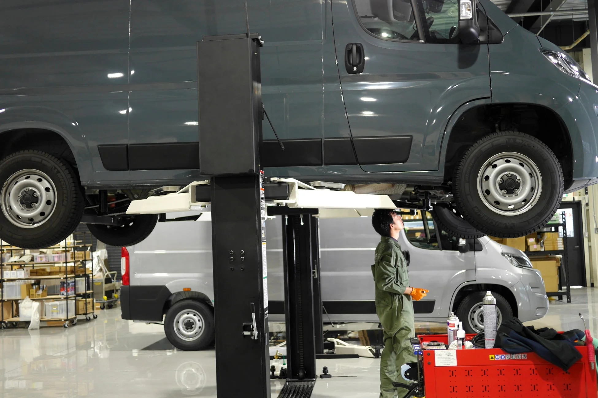 工場内で、作業員が自動車用リフトで高く持ち上げられたバンを下から点検しています。奥には別のバンも見え、整備作業が行われている様子です。