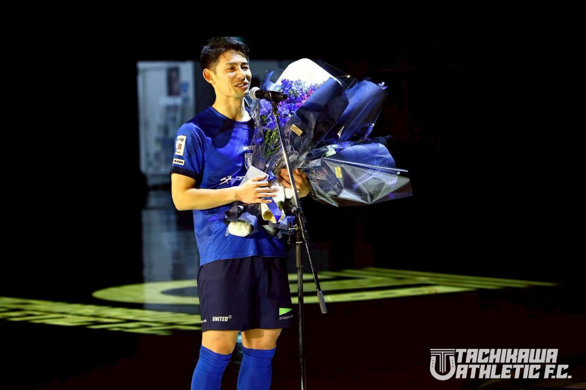 青いユニフォームを着た男性がステージ上で花束を受け取っている