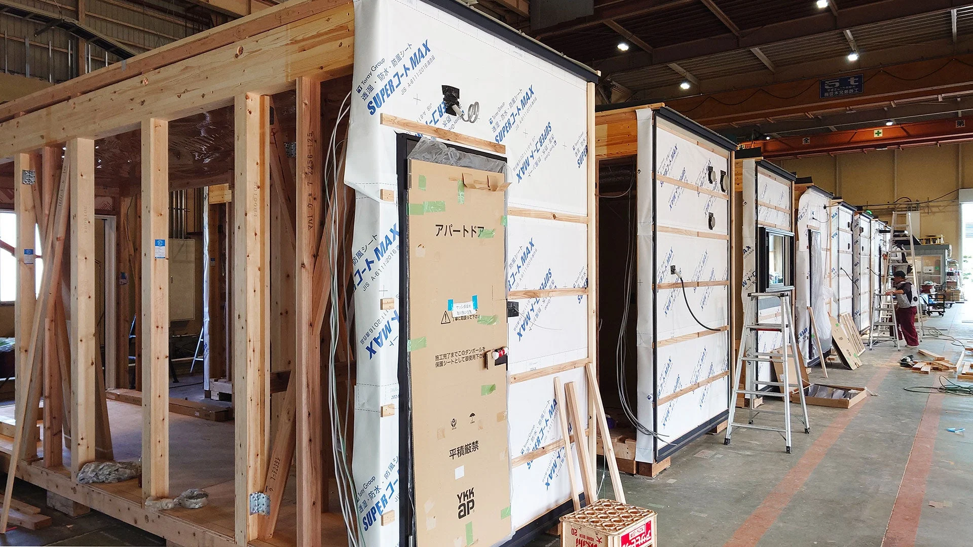 工場のような広い空間で、複数のプレハブ住宅やアパートの建設モジュールが並べられ、工事が進められている様子