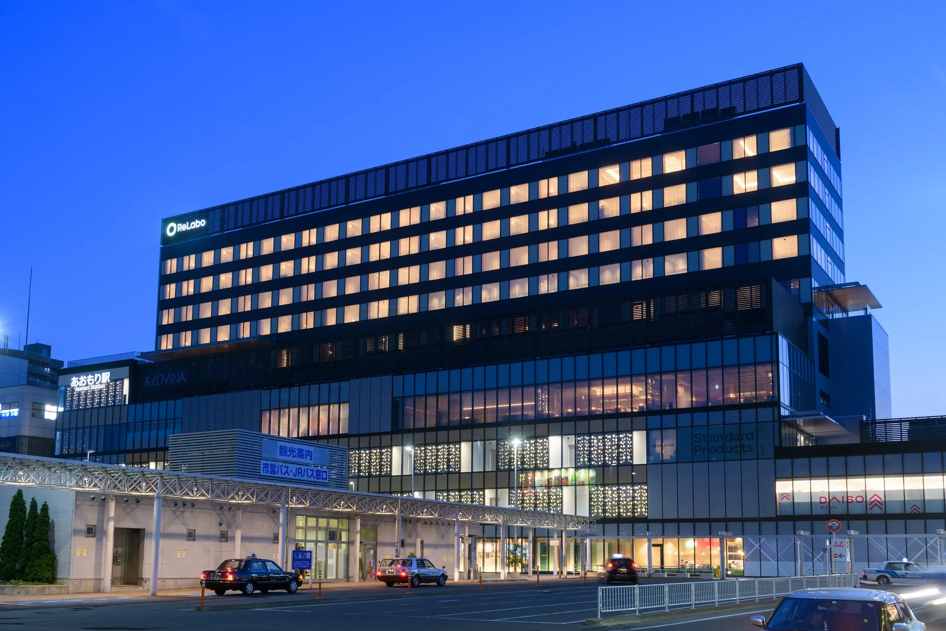 夕暮れ時または夜間にライトアップされたモダンな駅ビルと複合施設が写っています。手前にはバス・タクシー乗り場があり、活気ある都市の玄関口の様子がうかがえます。青森駅周辺の風景です。