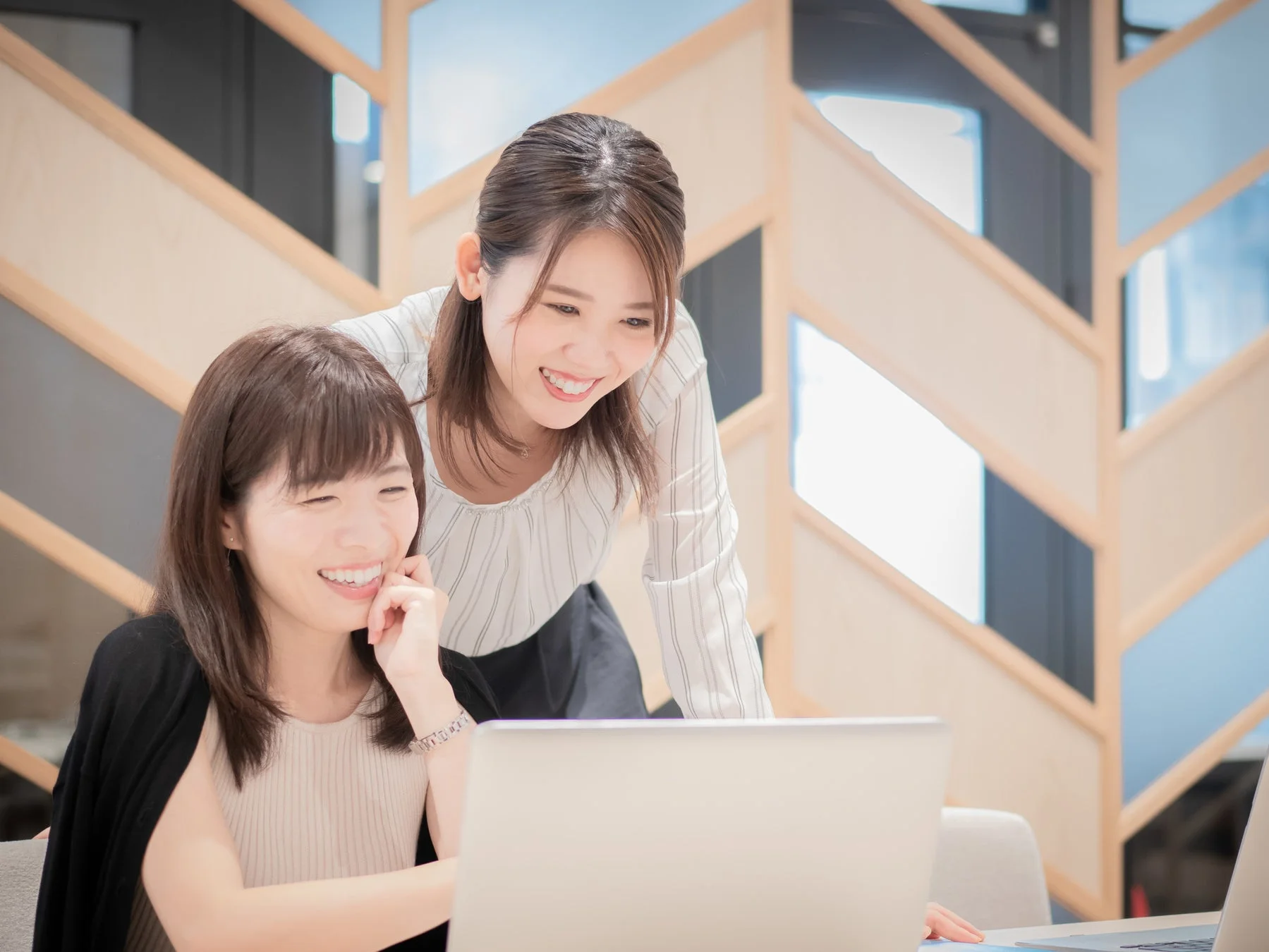 明るいオフィスで2人の女性がノートパソコンを見ながら笑顔で会話している様子。