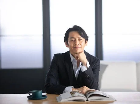 スーツを着てデスクに座り、カメラに微笑む中島輝氏