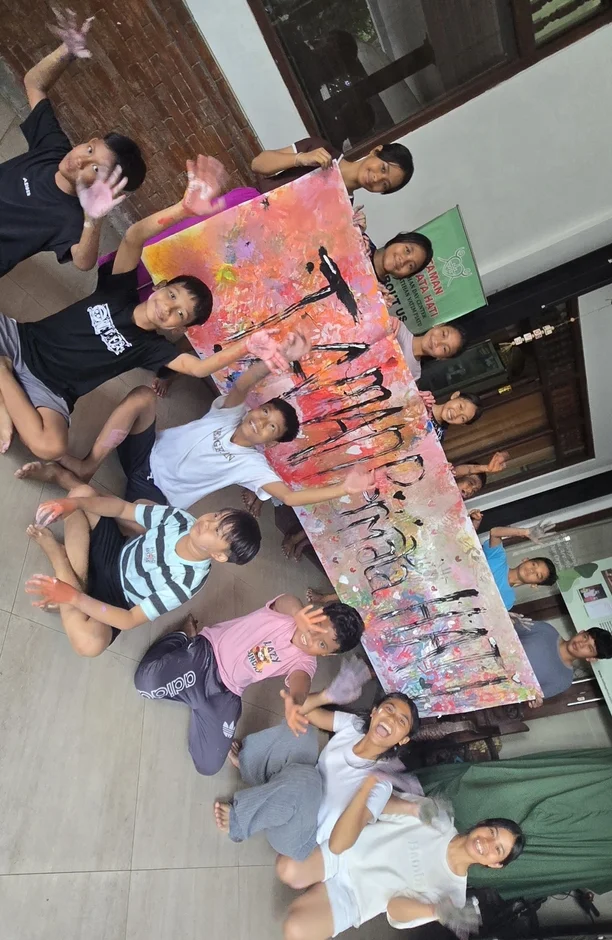 床に座った子供たちが、カラフルな手作りのバナーを笑顔で掲げています。バナーには「TAMAN HATI」と書かれ、子供たちの手には絵の具が付着しており、アート活動を楽しんだ様子が伺えます。背景には孤児院を示す看板も見えます。