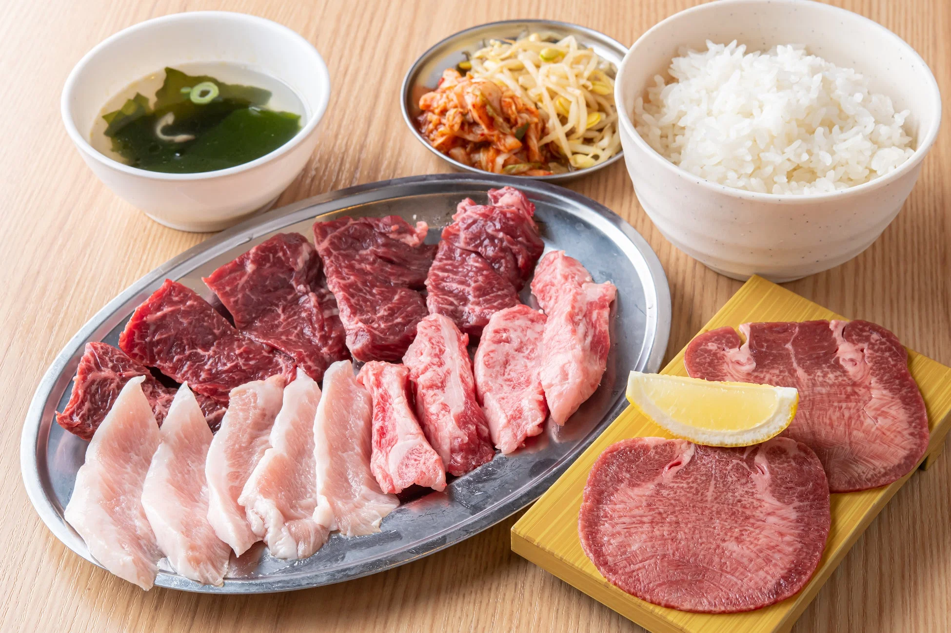 焼肉定食が並べられています。様々な部位の生牛肉と豚肉、レモンを添えた牛タン、白いご飯、わかめスープ、キムチと豆もやしのナムル