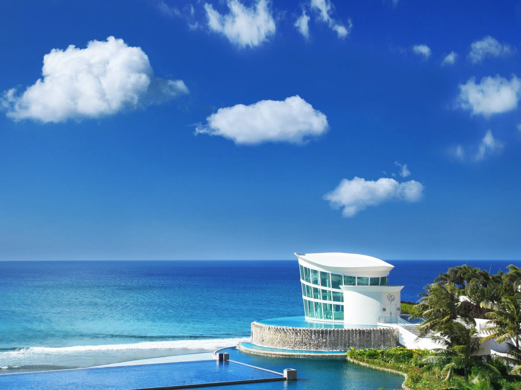 晴れた青空の下、海辺に建つモダンな白いチャペルとインフィニティプールが印象的なリゾート風景です。
