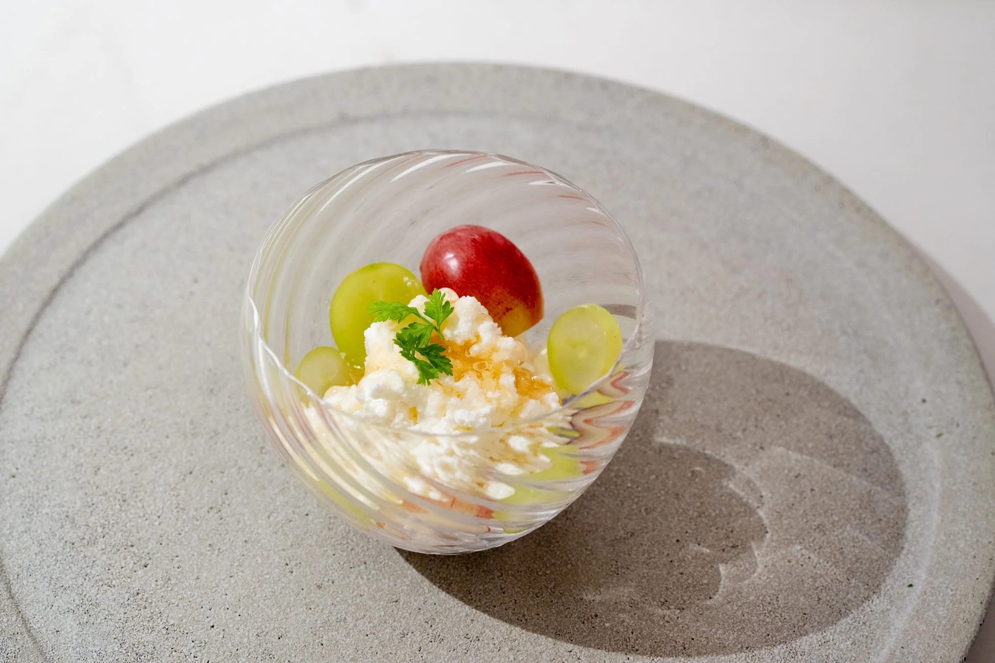 透明なグラスに入ったデザートで、白いクリーム状のものの上に緑と赤のぶどうが添えられ、はちみつがかかっています