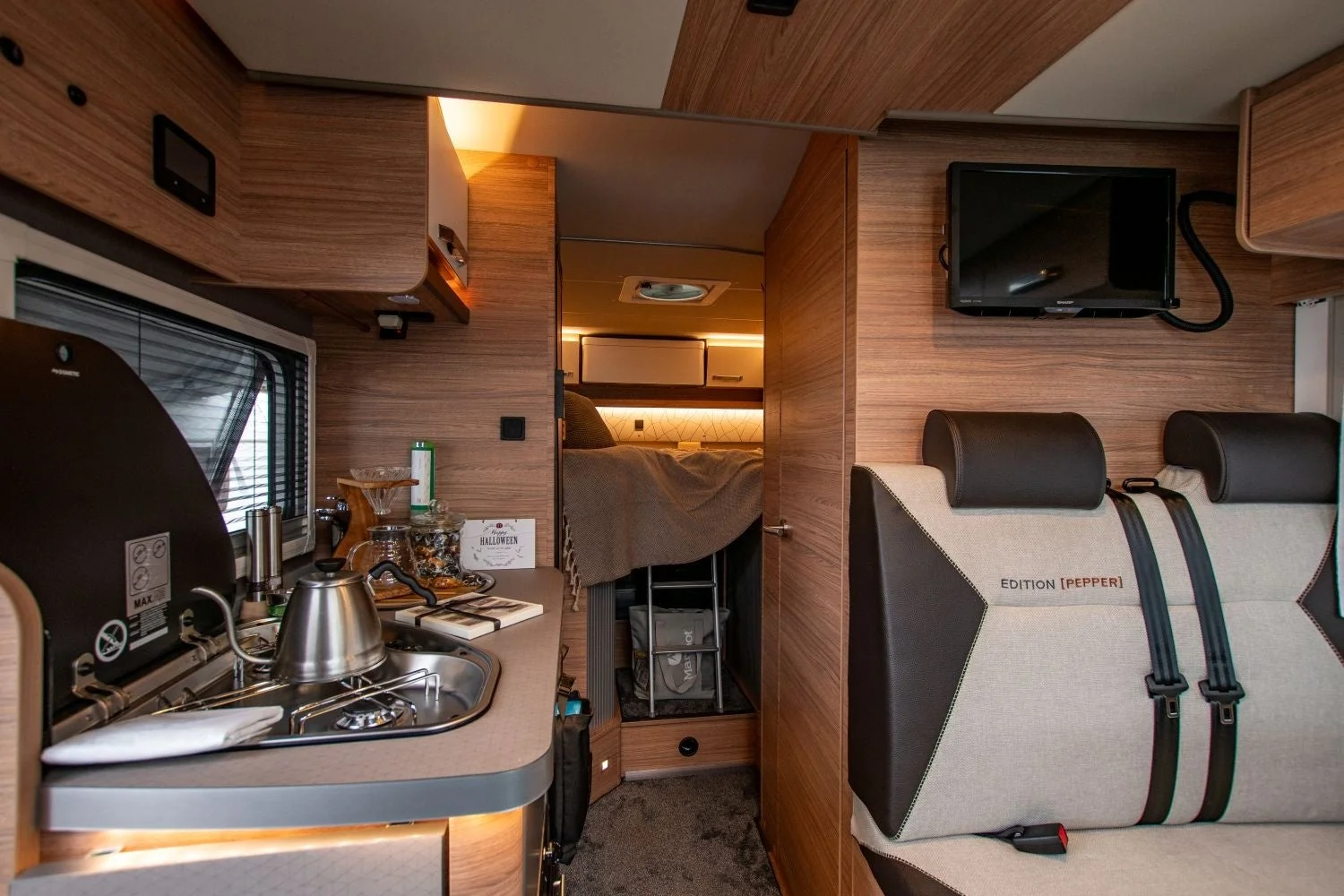 キャンピングカーの居心地の良い内装で、キッチン、リビング、寝室が一体となった空間が映し出されています。木目調の家具と温かい照明が特徴です。