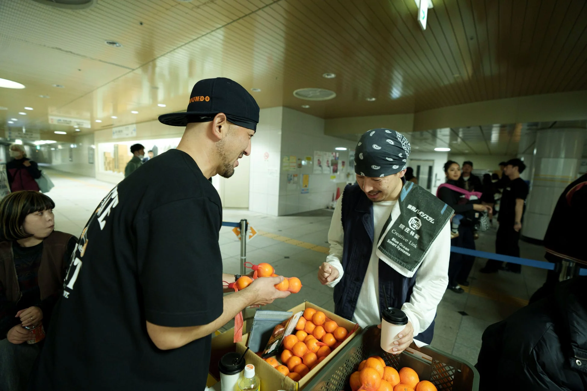 地下通路のような場所で、男性が別の男性にみかんを手渡している