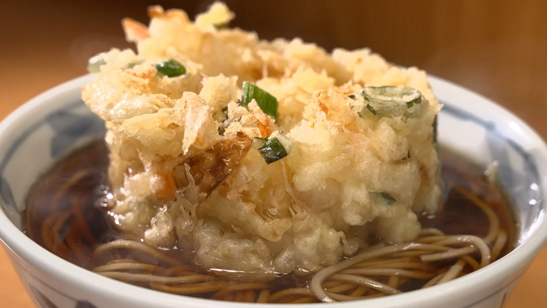 温かい出汁に浸されたそばの上に、サクサクとした衣のかき揚げが豪快に乗せられた一品。ネギの緑色が彩りを添えている。