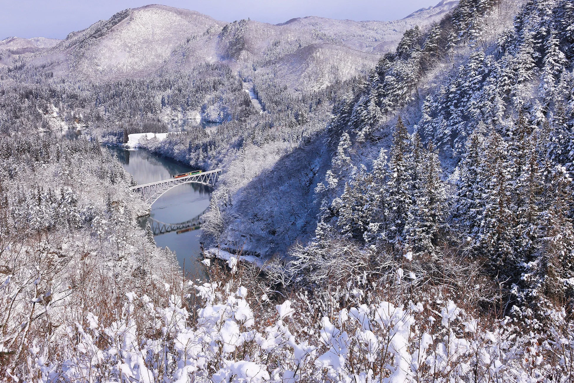 雪に覆われた雄大な山々と森林、川に架かる鉄橋を渡る列車