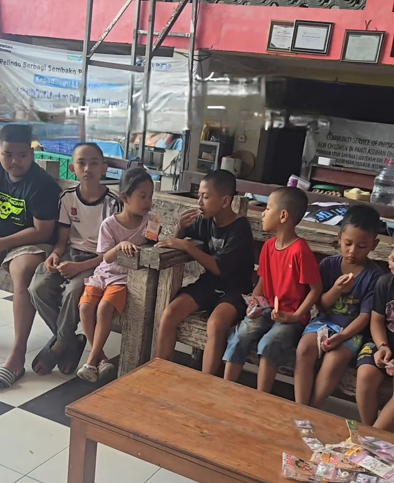 孤児院らしき場所で子供たちが木製のベンチに座り、お菓子を食べている様子。背景には社会奉仕活動に関するバナーが見える。