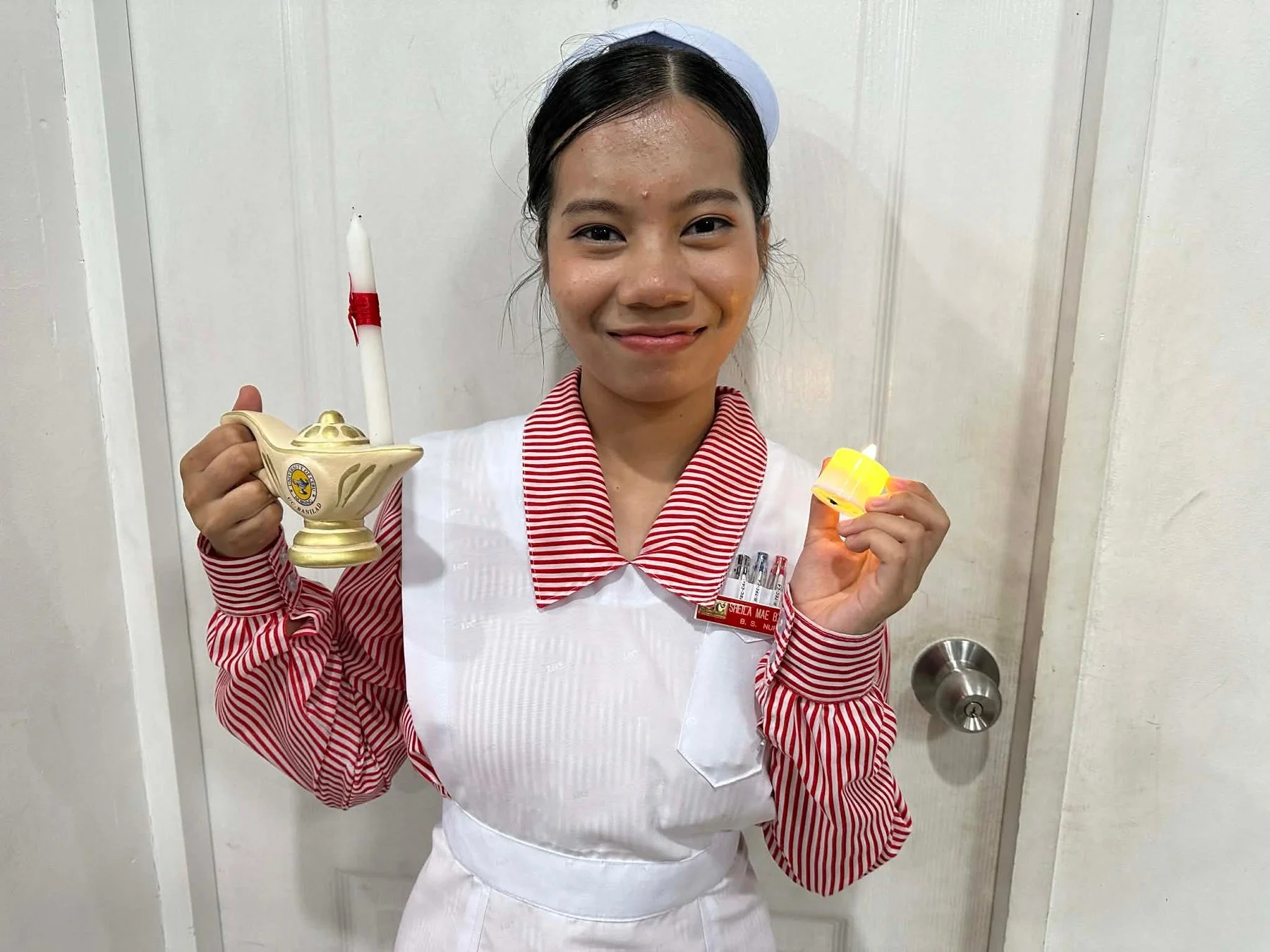 看護師の制服を着た女性が、ナイチンゲールランプとLEDキャンドルを持って笑顔で立っている。