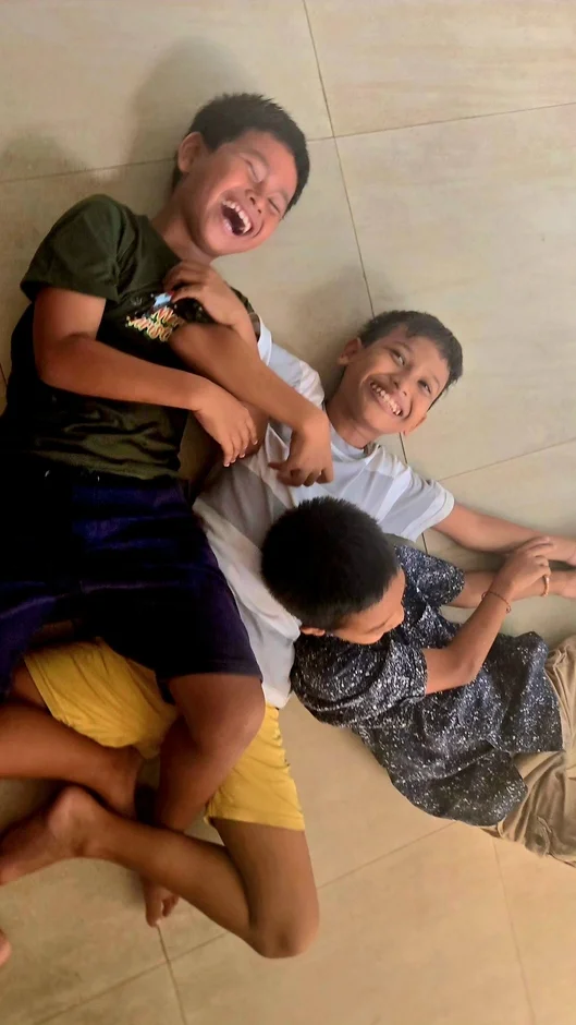 3人の子供たちが床に寝転がり、楽しそうに笑っている様子を捉えた画像です。無邪気な笑顔が印象的で、仲良く遊んでいる日常の一コマが表現されています。