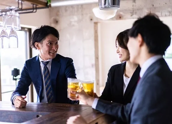 ビジネスウェアを着た男女3人がバーカウンターでビールグラスを手に笑顔で乾杯している様子