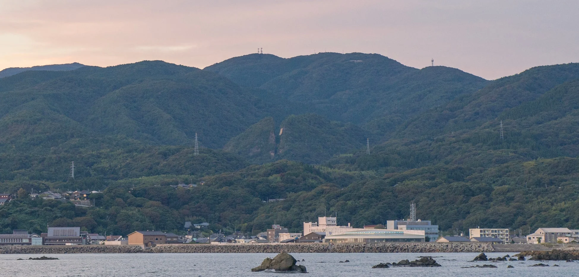 夕暮れ時の淡い空の下、緑豊かな山々を背景に、海岸線に沿って建物が立ち並ぶ町の風景