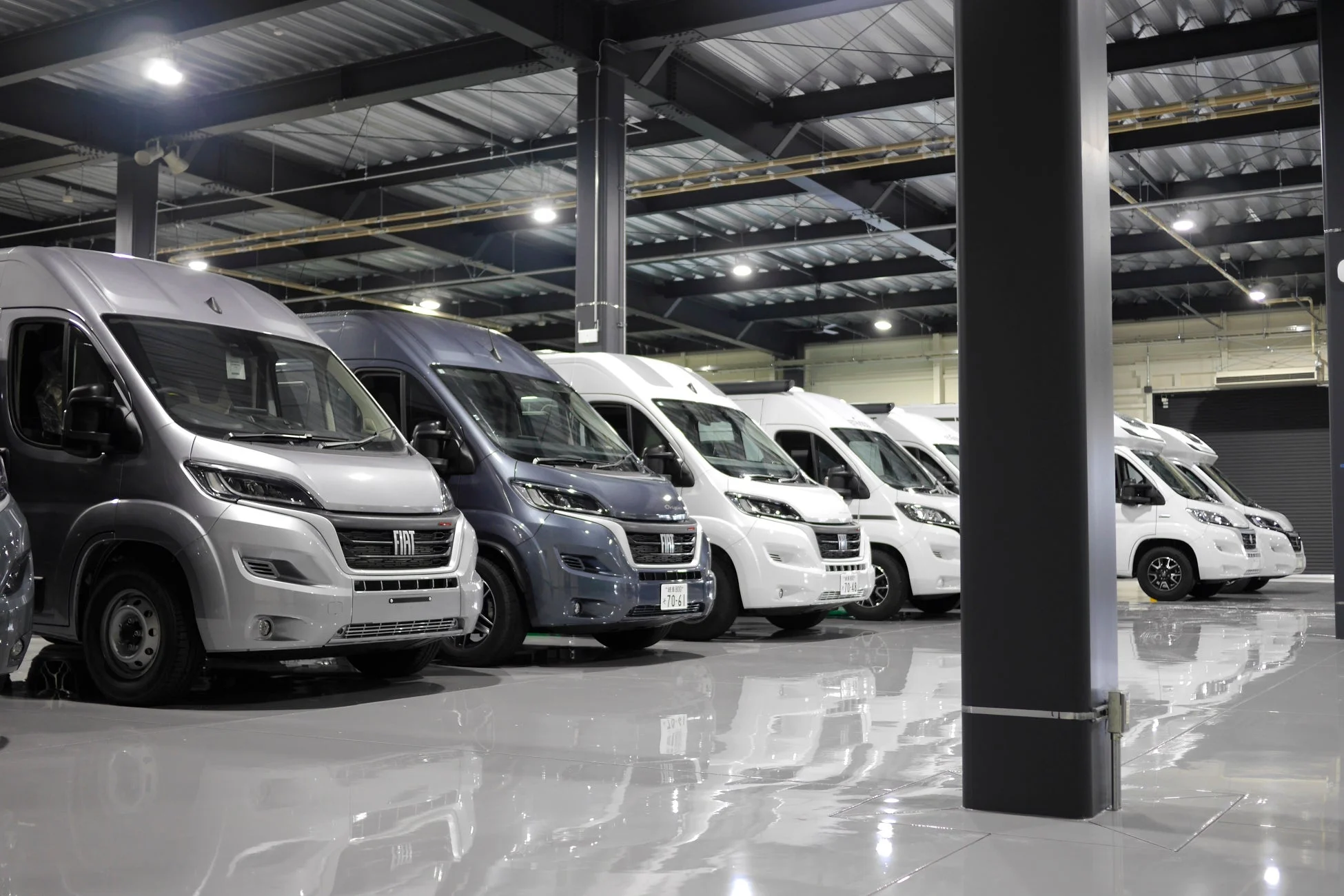 屋内施設に、フィアット・デュカトをベースとしたキャンピングカーや大型バンが複数台整然と駐車されています。床は光沢があり、車両が反射しています。