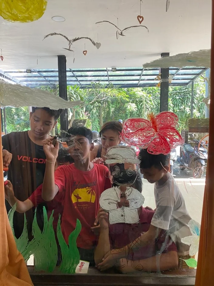 ガラス窓に絵を描く人々が写っています。子供から大人まで数人が集まり、太陽やハイビスカス、伝統的な人物の絵を共同で制作しているようです。背景には緑豊かな屋外の景色が見え、楽しげな創作活動の様子が伝わります。