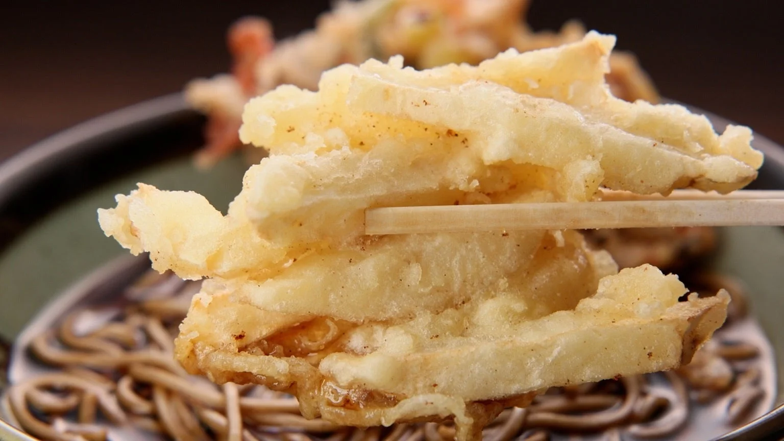 箸で持ち上げられたサクサクのかき揚げが、温かい蕎麦の上で輝いています。揚げたての美味しさが伝わる、食欲をそそる日本の伝統料理の瞬間を捉えた一枚です。