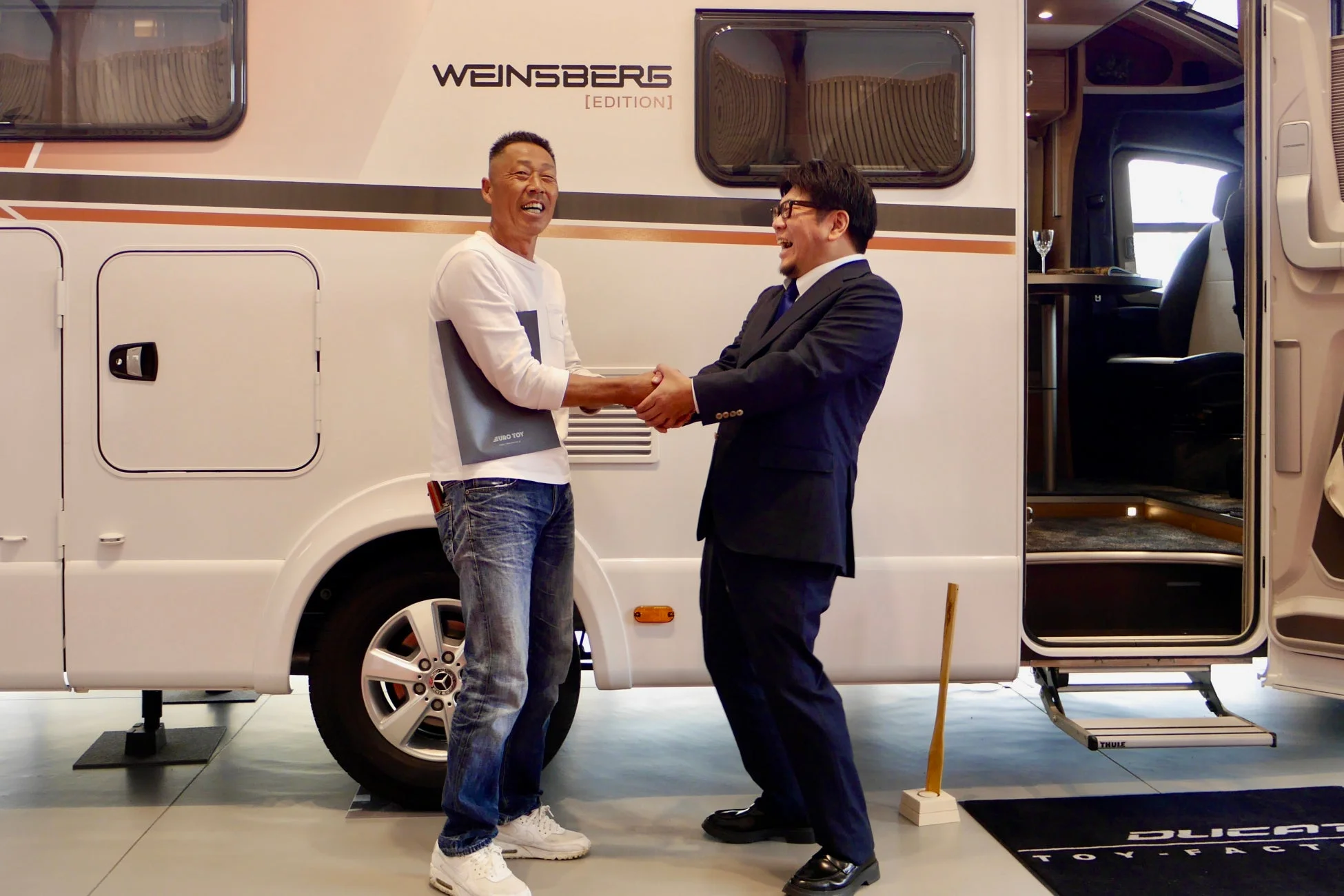 白いキャンピングカーの前で、二人の男性が笑顔で握手を交わしています。一人は「EURO TOY」と書かれたファイルを持ち、キャンピングカーには「WEINSBERG [EDITION]」の文字が見えます。展示会か商談の場で喜びを分かち合っている様子です。