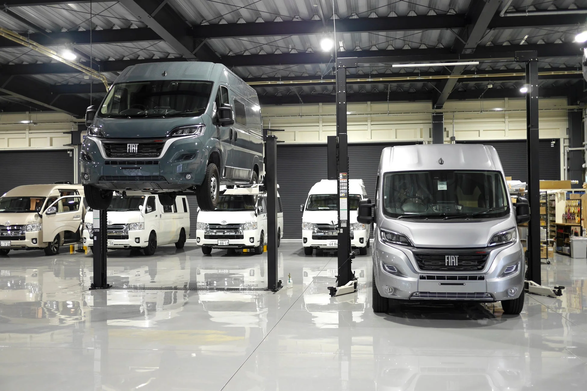 広々とした整備工場で、複数のバンが整備されている様子です。中央にはグレーのフィアット車がリフトで持ち上げられ、手前にはシルバーのフィアット車、奥には白いトヨタハイエースが複数台並んでいます。床は光沢があり、清潔感のある作業場です。