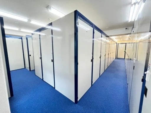 青いカーペットが敷かれた明るい室内空間に、白い扉と濃い青色のフレームを持つ多数の貸し倉庫（トランクルーム）が整然と並ぶ廊下の様子