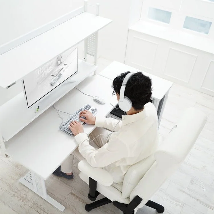 白い部屋でヘッドホンを着用した男性が白いデスクと椅子に座り、パソコンで作業している様子。全体的に白で統一されたミニマリストな空間で、モニターには白い製品の画像が表示されている。