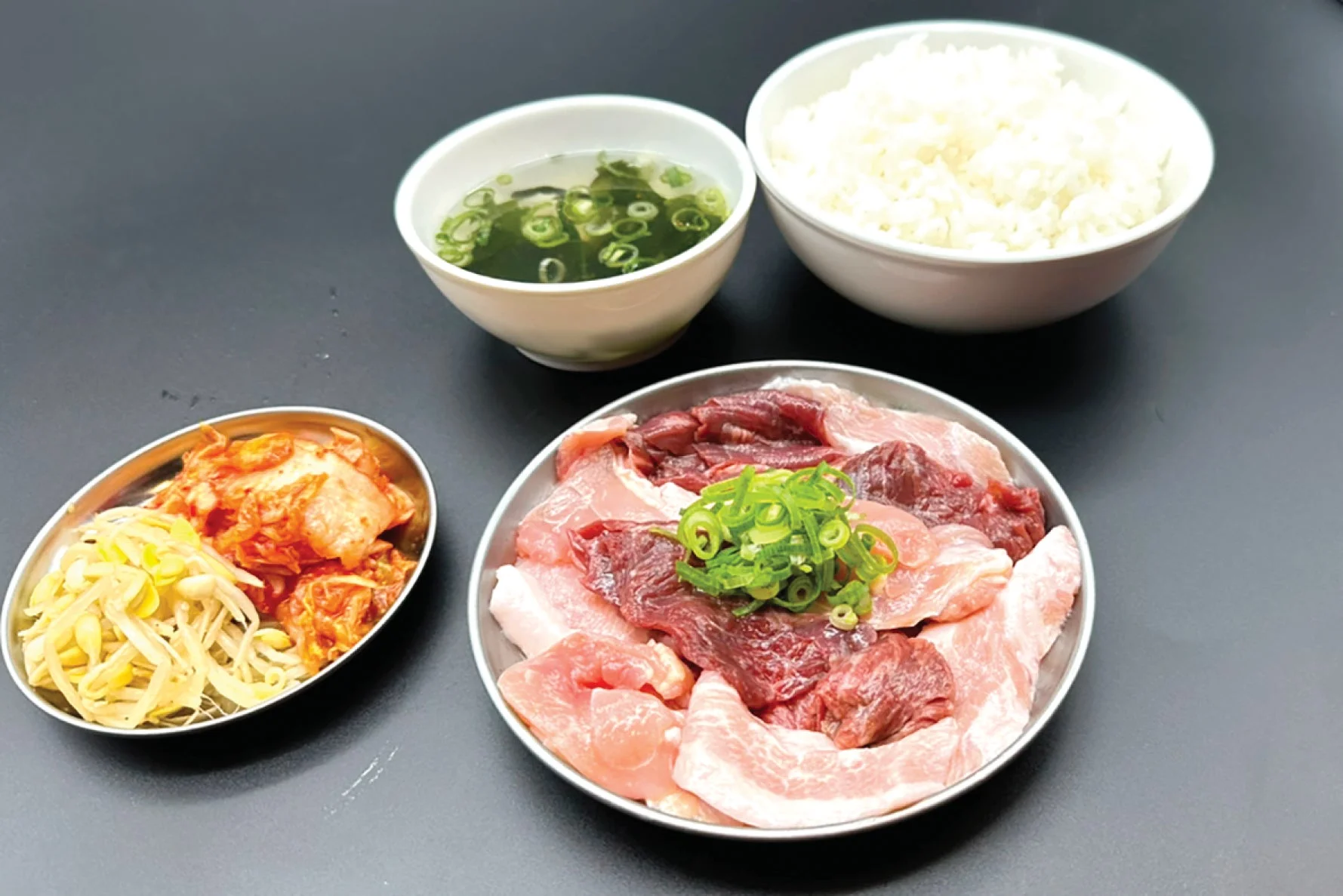 焼肉用の様々な種類の生肉が盛られた皿を中心に、ご飯、ワカメスープ、キムチと豆もやしのナムルが並べられた食事のセット