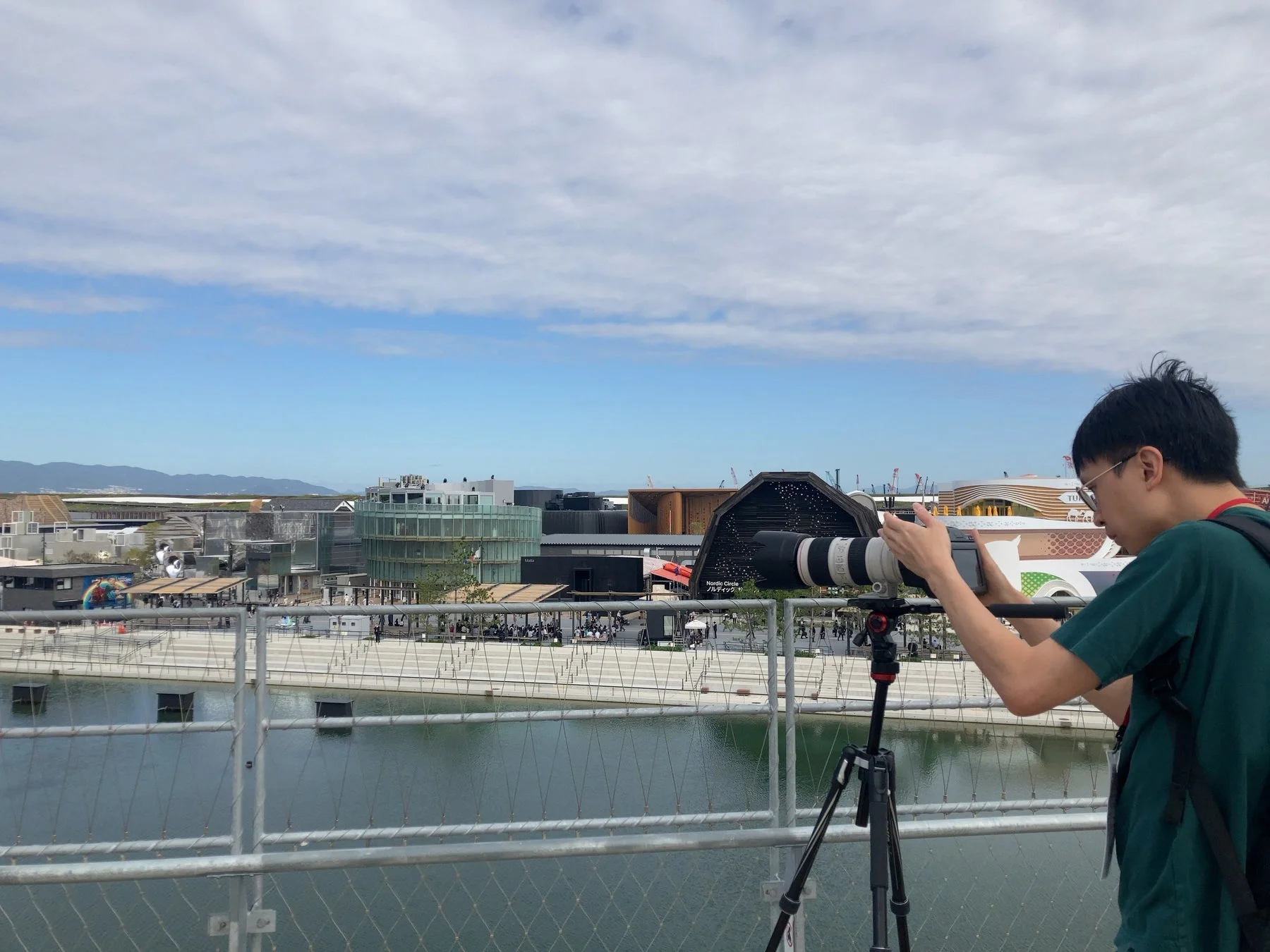 男性が三脚に据え付けた望遠カメラで、水辺に広がるモダンな建物群を撮影している