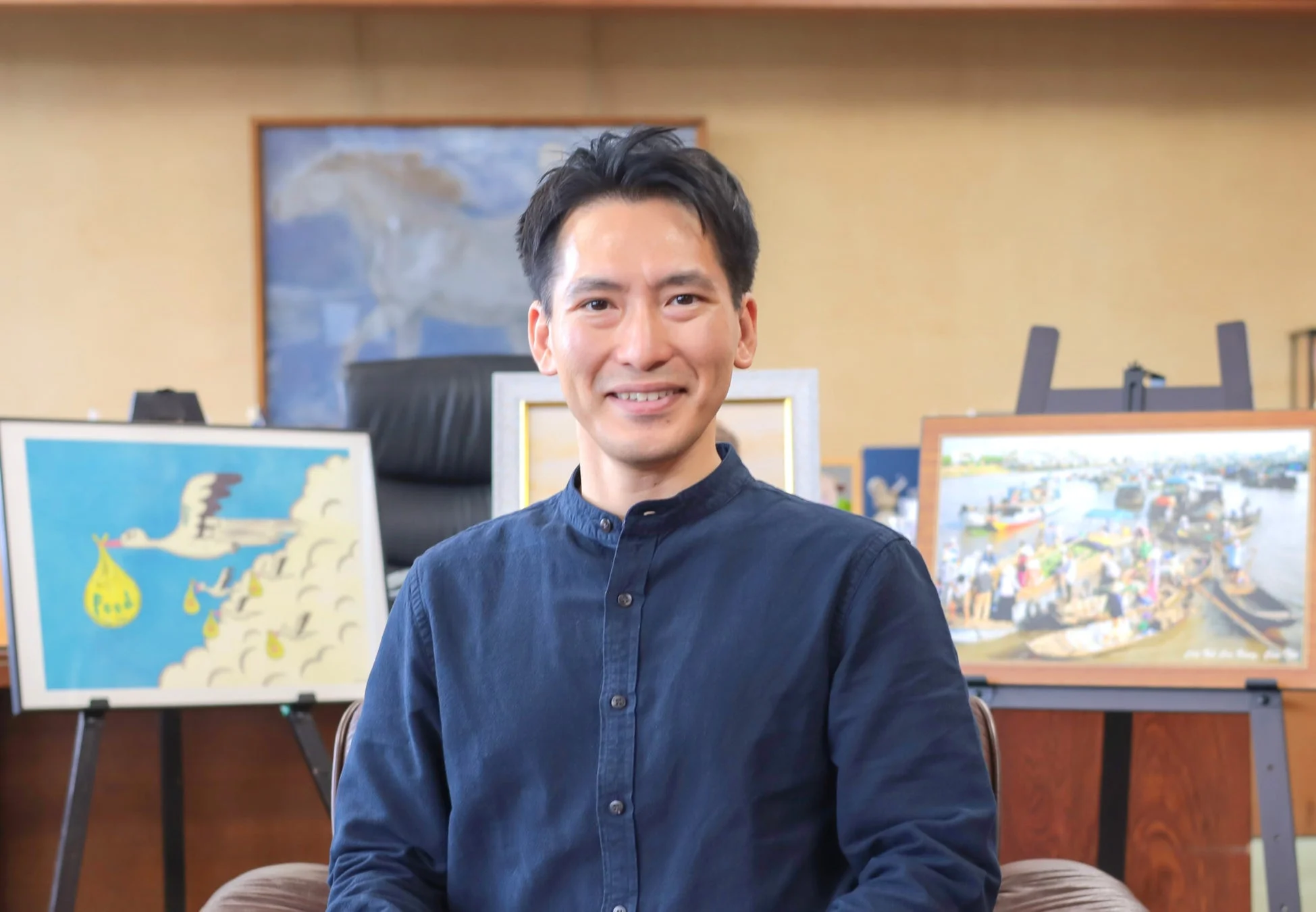 紺色のシャツを着た男性が笑顔でこちらを見ています。背景には、コウノトリが何かを運ぶ絵や、活気ある水上市場のような絵が飾られています。