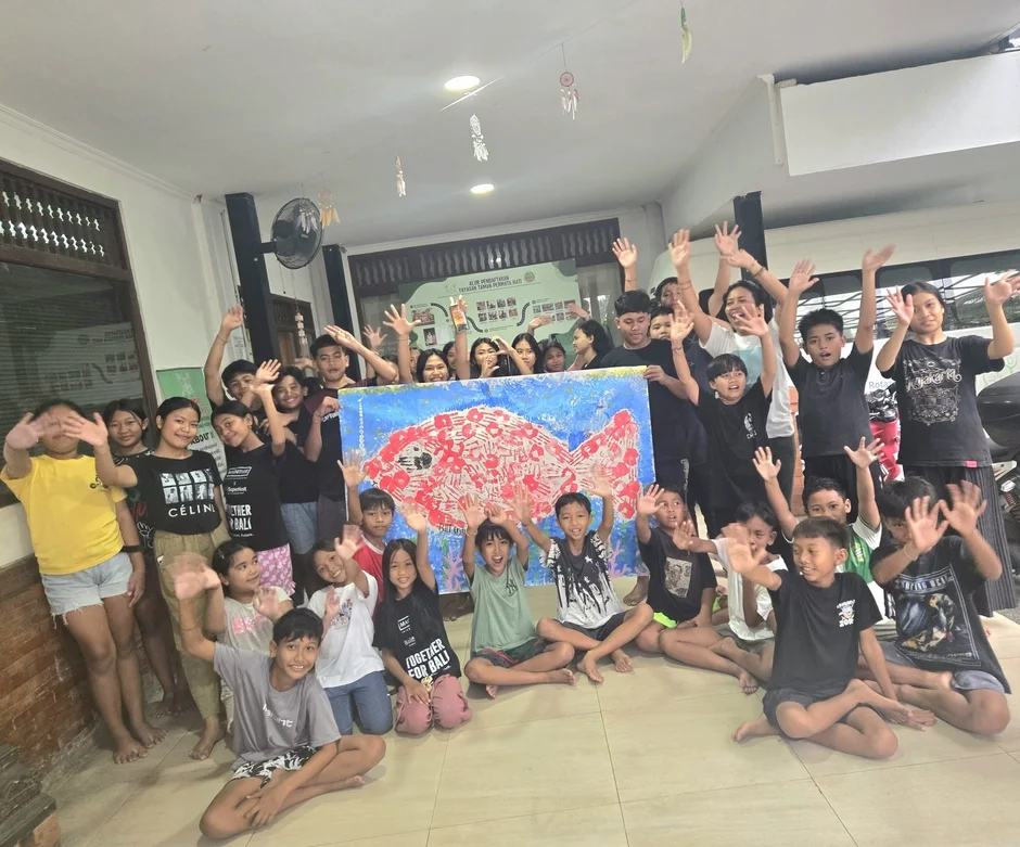 多くの子供たちと数人の大人が、ハンドプリントで描かれた赤い魚のアート作品を囲んで、手を振って笑顔でポーズをとっている集合写真です。楽しげなコミュニティ活動の様子が伝わります。