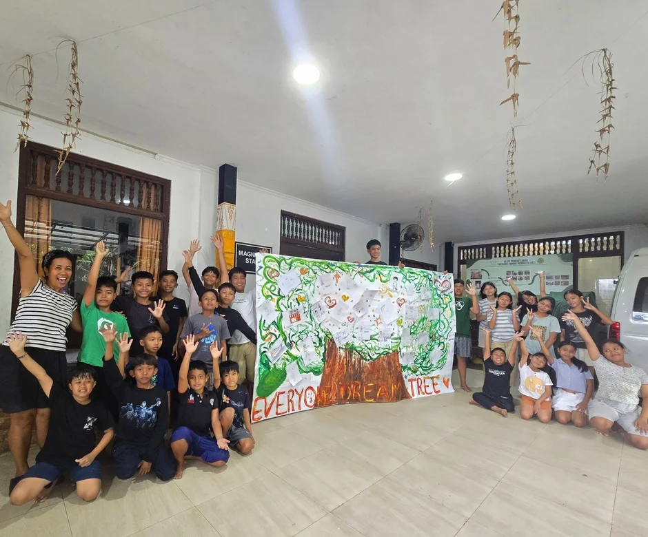 屋内イベントで、子どもたちと大人が「EVERYONE'S DREAM TREE」と書かれた大きなアートポスターを囲んで笑顔でポーズ。皆で協力して作品を完成させた喜びが伝わる、活気ある一枚です。