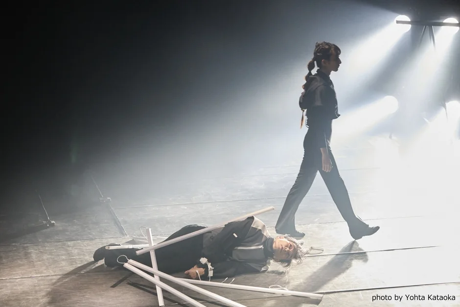 煙が立ち込めるステージで、一人が歩き、もう一人が床に倒れている様子を捉えた写真。強いスポットライトが当たり、幻想的でミステリアスな雰囲気を醸し出している。