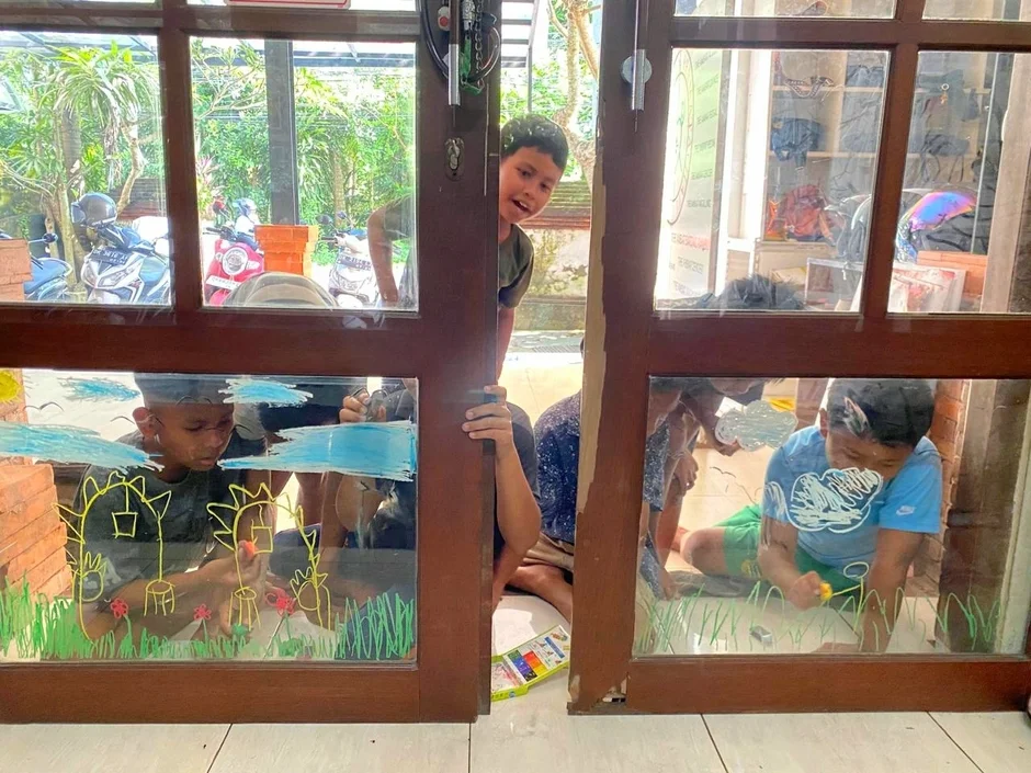 複数の子供たちが屋内の窓ガラスに絵を描いて遊んでいます。一人の子供が外を見て笑顔を見せており、楽しそうな創作活動の様子が伝わります。